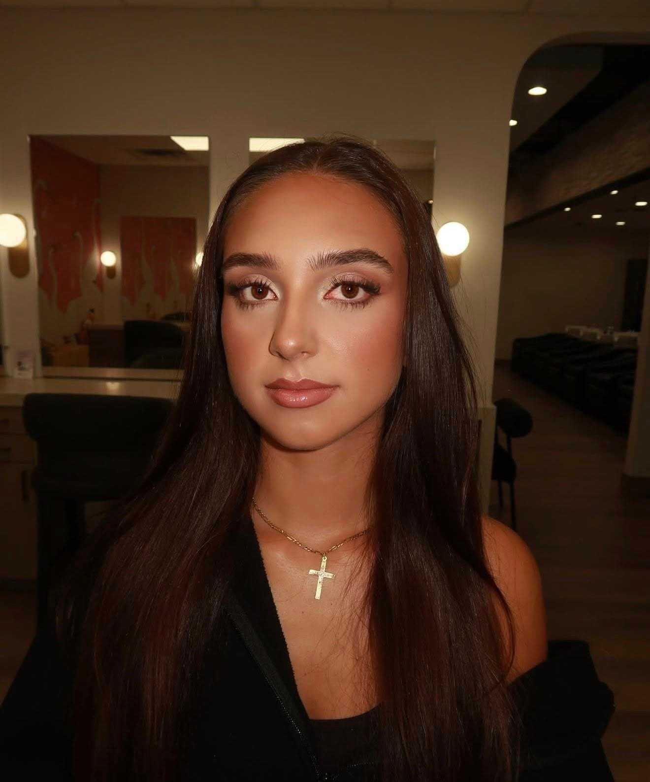 Young woman with long brown hair in a dimly lit room, wearing a gold cross necklace.